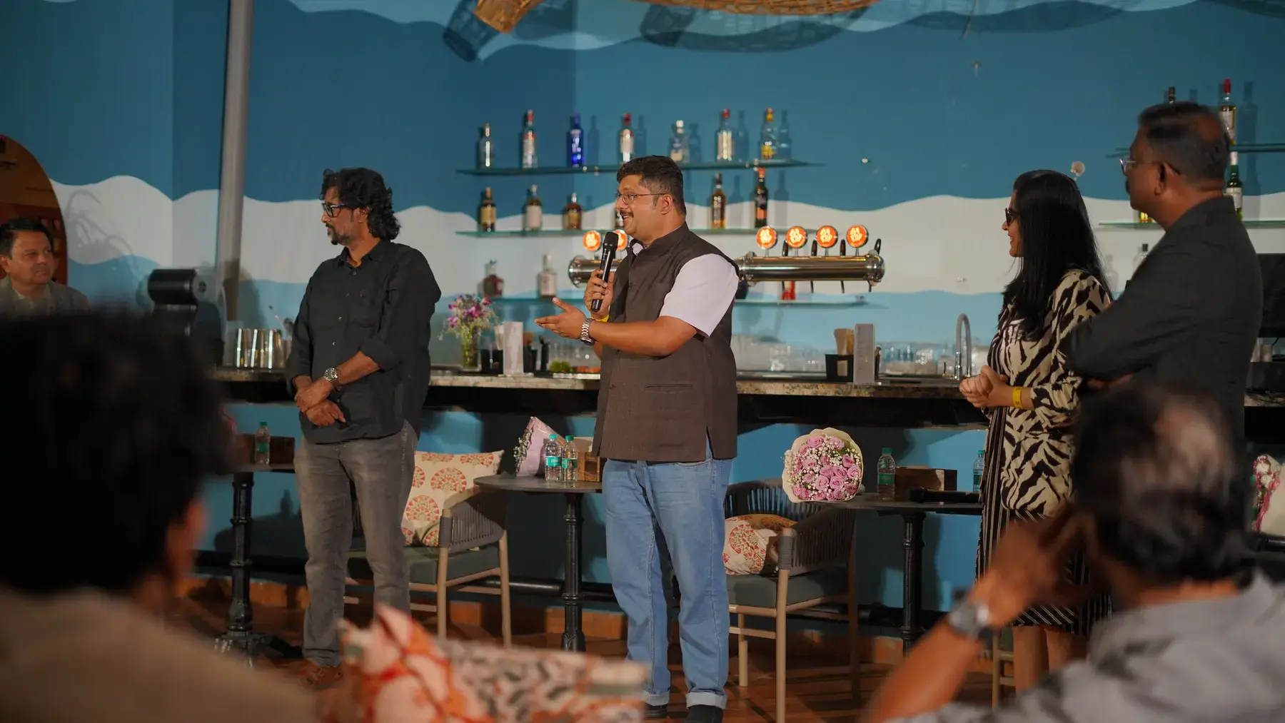 A speaker addressing the seated room with a microphone, standing in front of the bar